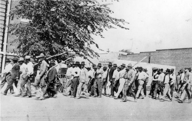 Eine Gruppe von Truppen der Nationalgarde, die Gewehre mit angebrachten Bajonetten tragen, eskortieren unbewaffnete schwarze Männer nach dem Massaker von Tulsa Race, Tulsa, Oklahoma, Juni 1921, in ein Internierungslager.