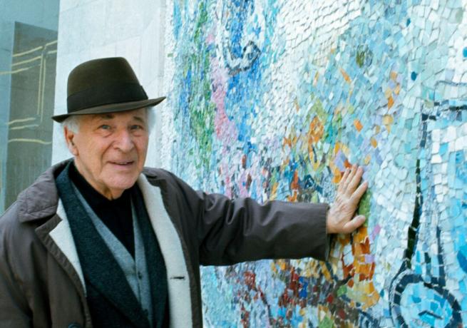 Der Künstler Marc Chagall, der einen Hut trägt, drückt seine Hand gegen eine Wand mit blauen Mosaikmustern.