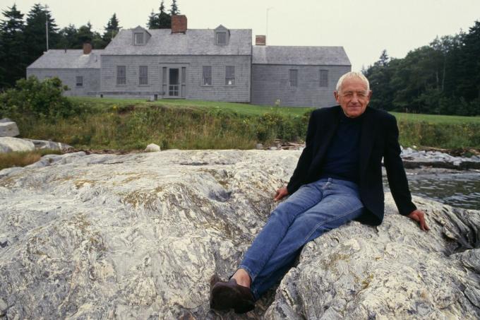 weißhaariger Mann, der auf Felsen vor einem grau gestalteten horizontal ausgerichteten Haus in Neuengland sitzt