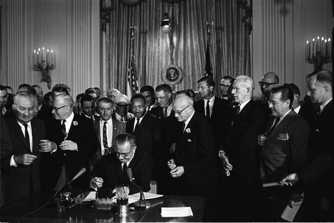 Präsident Lyndon B. Johnson unterzeichnet das Bürgerrechtsgesetz von 1964, während Martin Luther King, Jr. und andere zusehen.