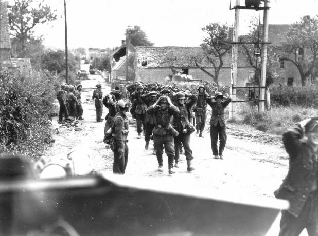 Deutsche Soldaten marschieren kapitulierend mit den Händen auf dem Kopf eine Straße entlang.